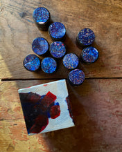 Blue glittery lidded amber jars on a wooden surface with a small painting below.
